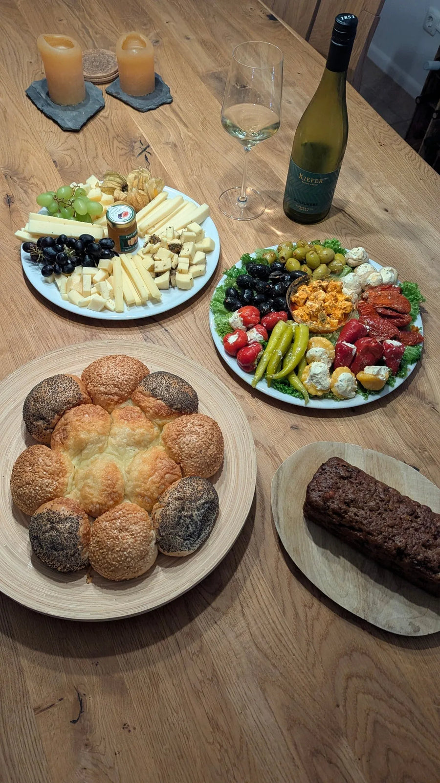 Käseplatte, Antipasti und frisches Brot auf einem Holztisch, daneben Weißwein im Glas