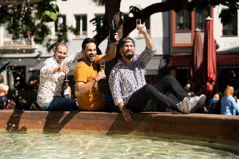 3 Männer genießen Sekt im Freien an einem Brunnen